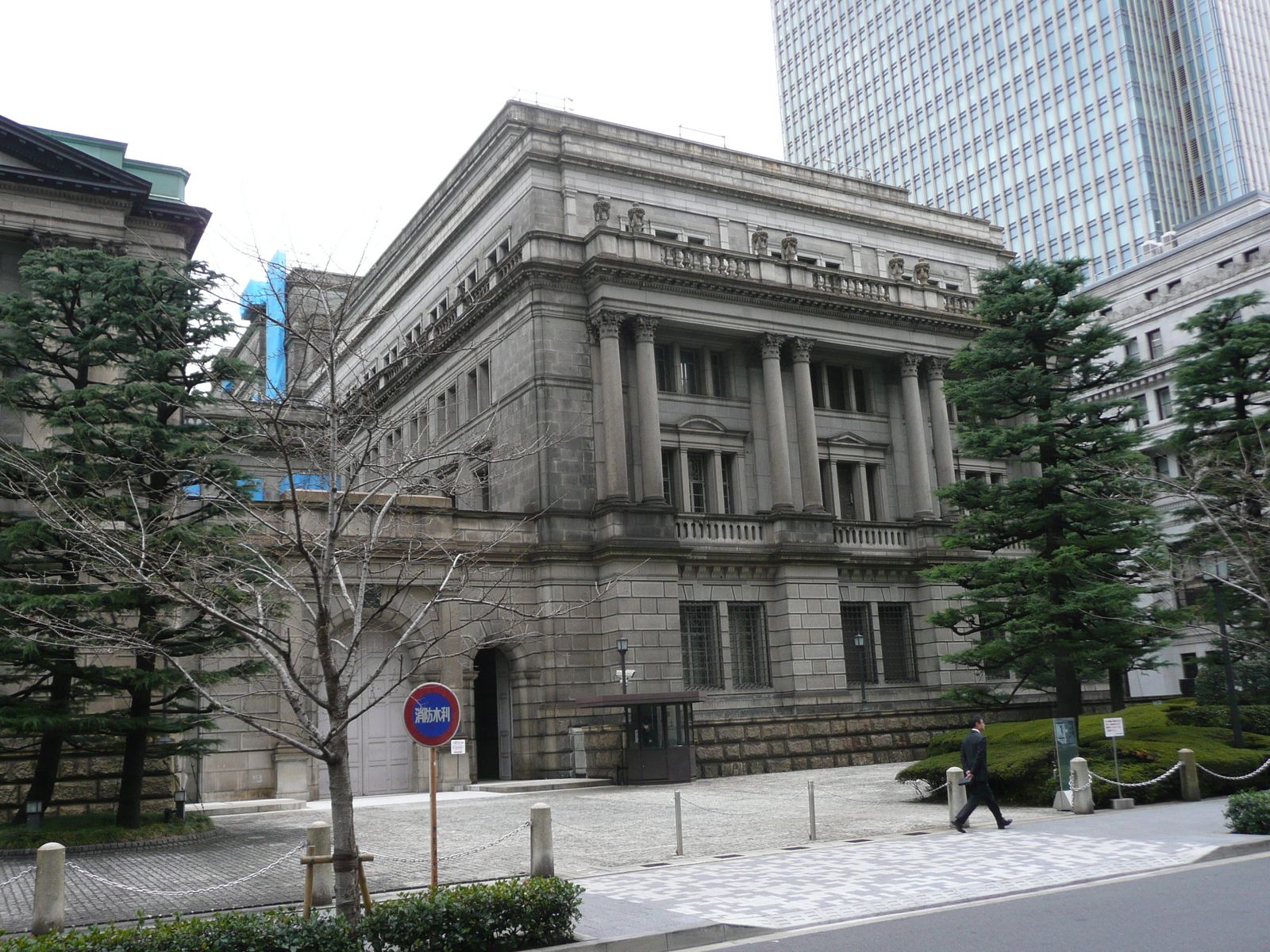 The stone facade of the Bank of Japan head office in Tokyo, with pine trees in the foreground and a glass office tower behind